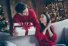 Nápad na vánoční dárek pro ni - muž dává dárek ženě, oba mají na sobě červené svetry, kolem sváteční atmosféra
