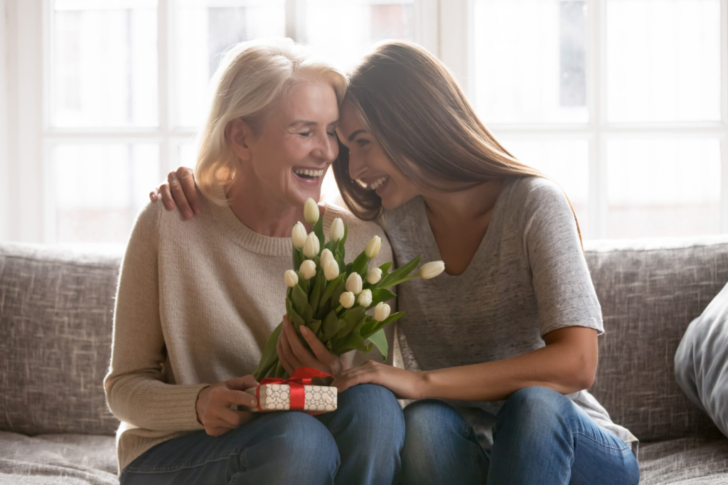 Dvě ženy, které sedí na sedačce a usmívají se. Jedna žena v ruce drží květiny a dárek.