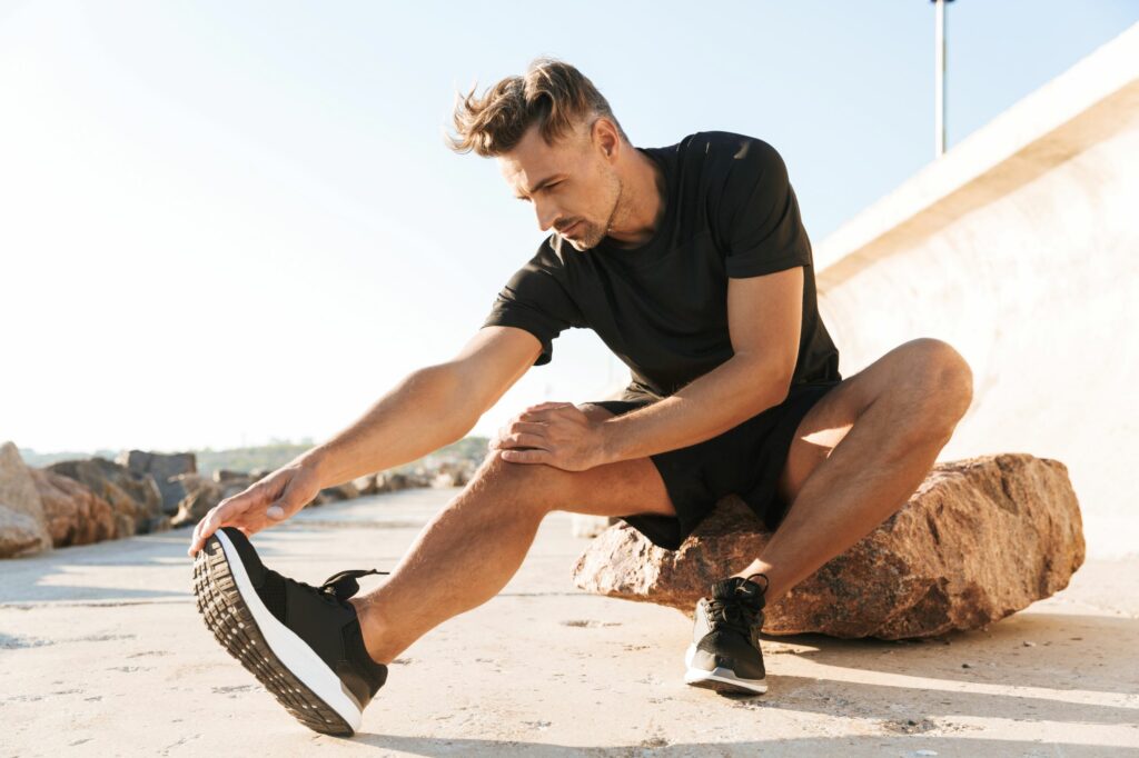 Muž protahující se před ranním joggingem na pláži v černém tričku, šortkách a pánské sportovní obuvi
